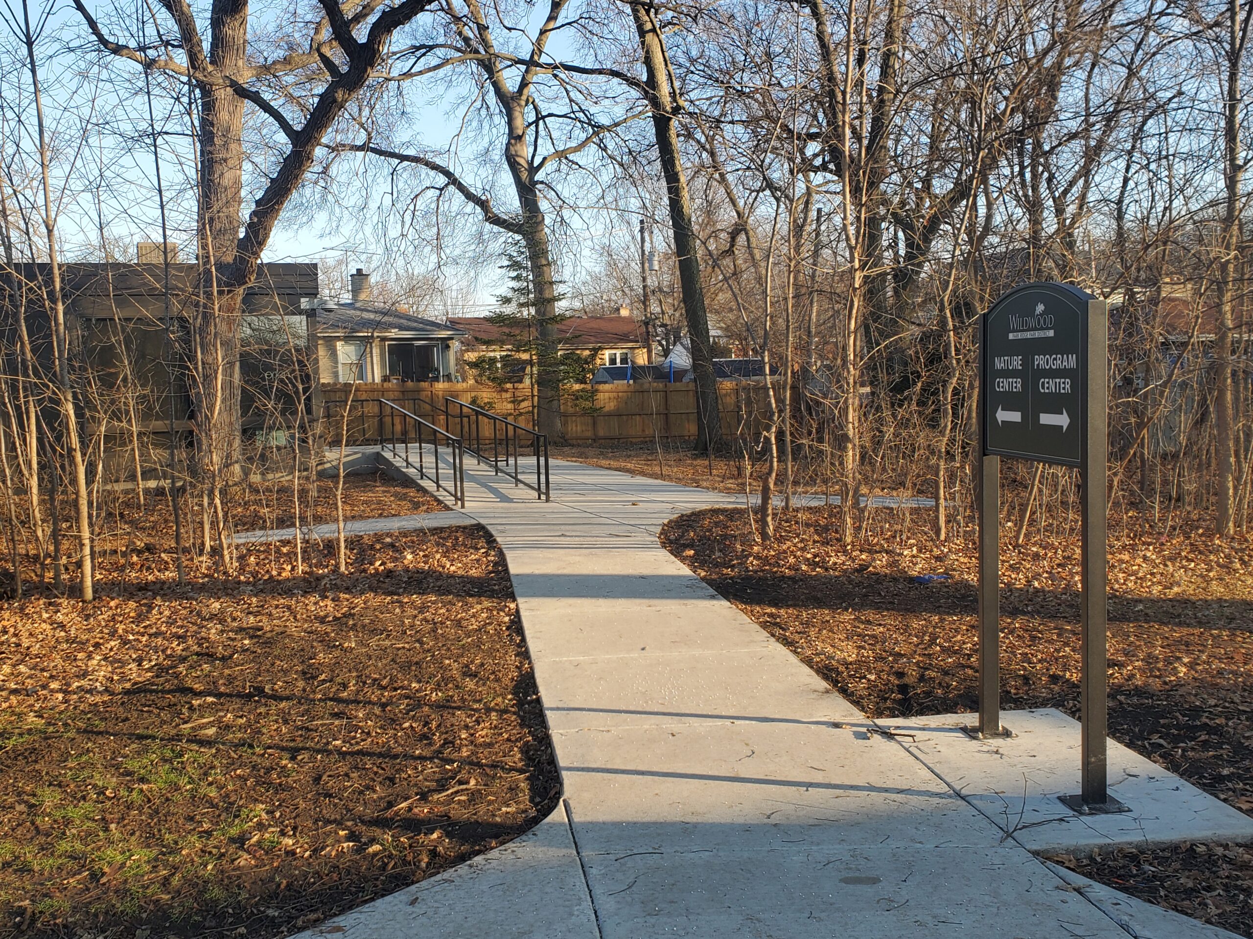 a poured concrete walkway and ramp leading through a yard and trees to the Wildwood Nature Center separate building